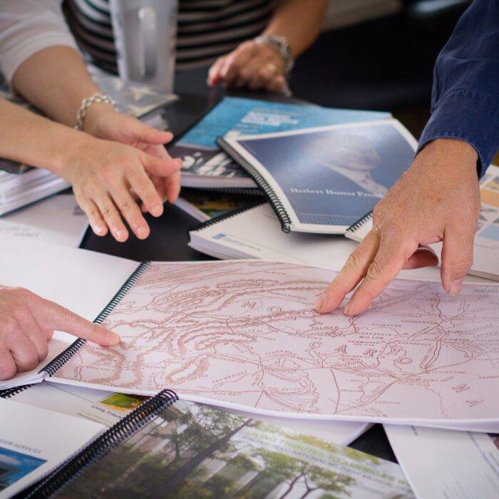 Close-up of hands pointing at a spread of exhibit design materials and a hand-drawn map across a worktable, Eisterhold Associates studio.