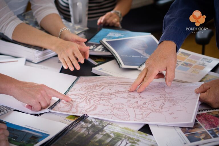 Close-up of hands pointing at a spread of exhibit design materials and a hand-drawn map across a worktable, Eisterhold Associates studio.