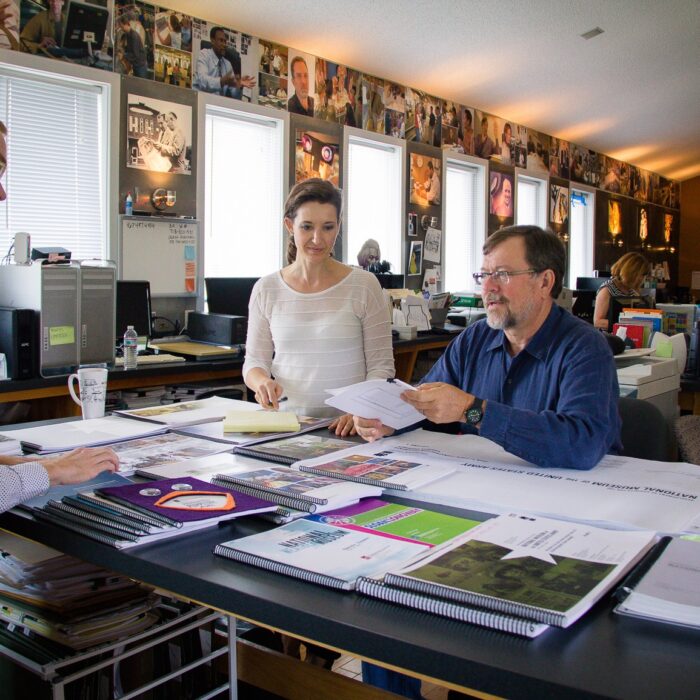 Team of five gathered around a worktable reviewing exhibit design proofs and materials, Eisterhold Associates studio, museum photography lining the walls behind them.