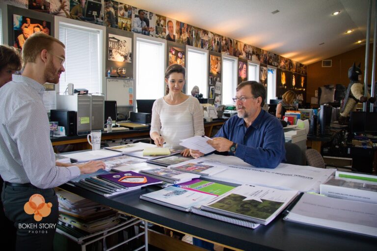 Team of five gathered around a worktable reviewing exhibit design proofs and materials, Eisterhold Associates studio, museum photography lining the walls behind them.