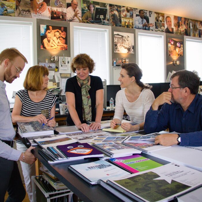 Team of five gathered around a worktable reviewing exhibit design proofs and materials, Eisterhold Associates studio, museum photography lining the walls behind them.