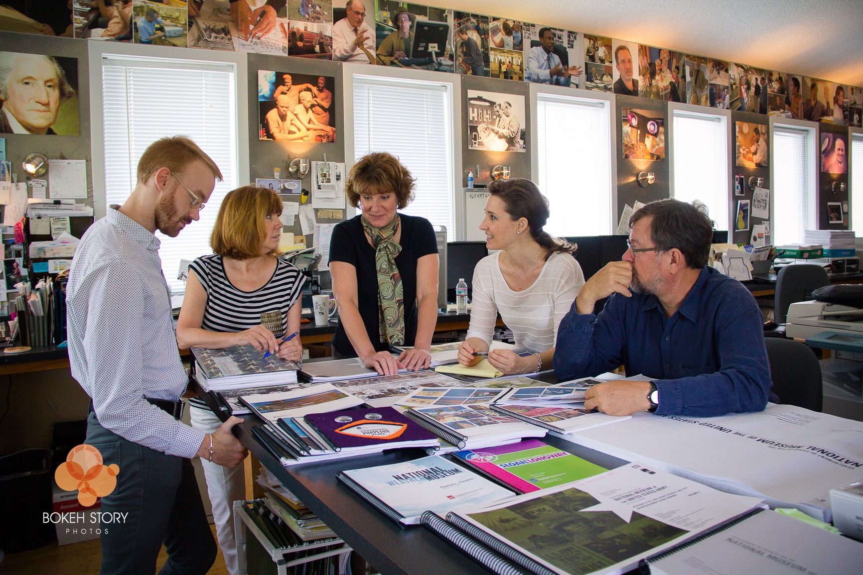 Team of five gathered around a worktable reviewing exhibit design proofs and materials, Eisterhold Associates studio, museum photography lining the walls behind them.
