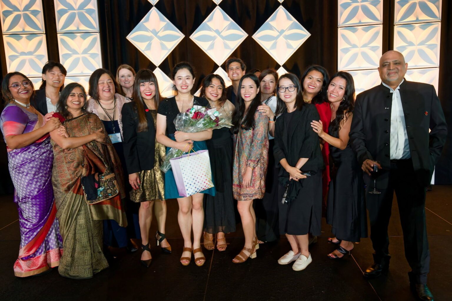 Evening gala group photo in front of a lit geometric backdrop, one attendee holding flowers and a gift bag, Leadership Convention, Kansas City, August 2025.