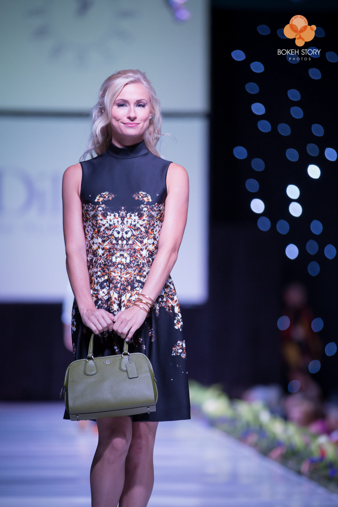 Runway photo of a model in a black sleeveless dress with an abstract floral print, carrying an olive green handbag, at the Adorn Style Show.