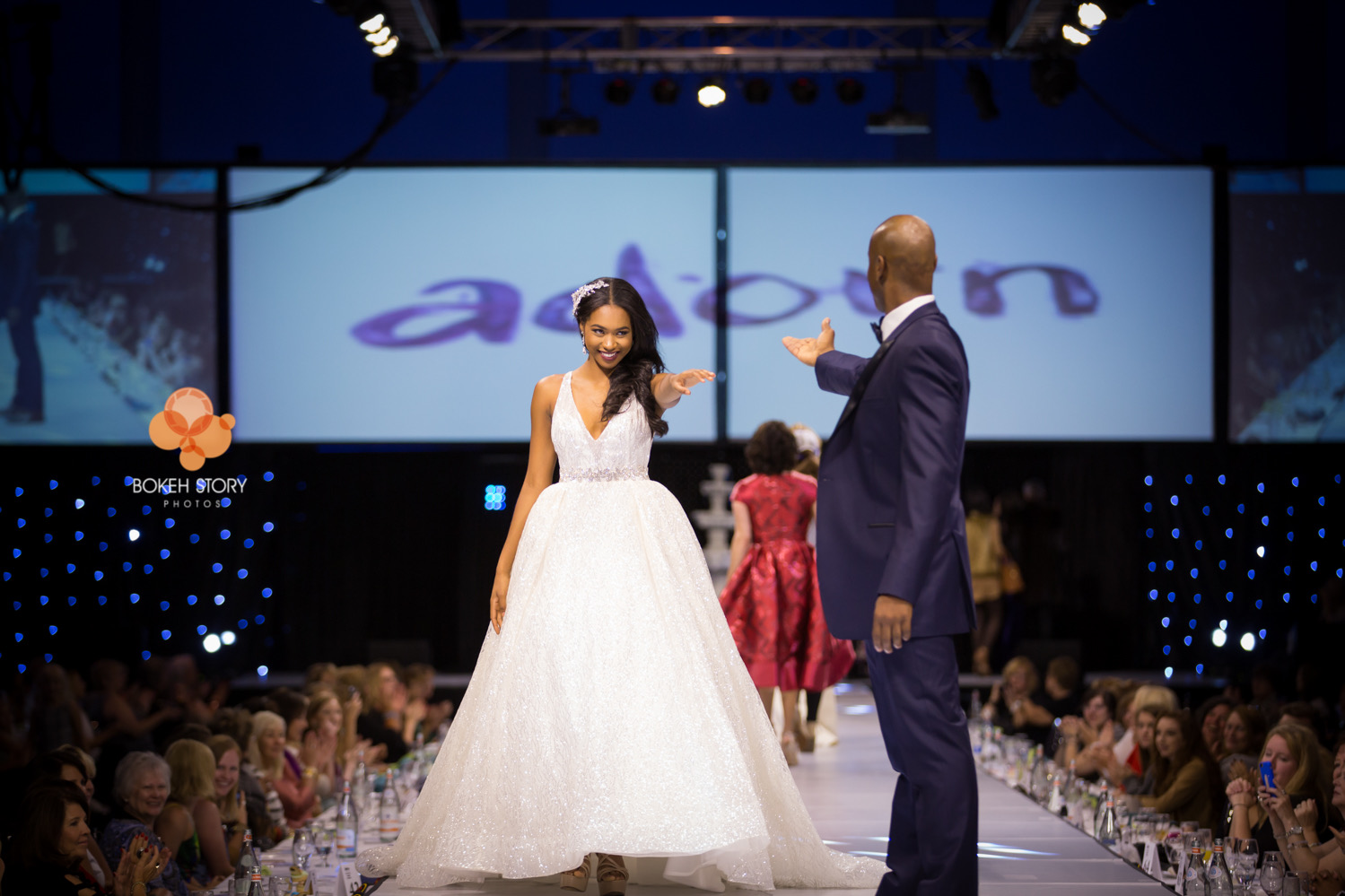 Runway photo of a model in a white beaded wedding gown reaching toward an emcee in a navy tuxedo, with the Adorn show logo projected on screens behind them.