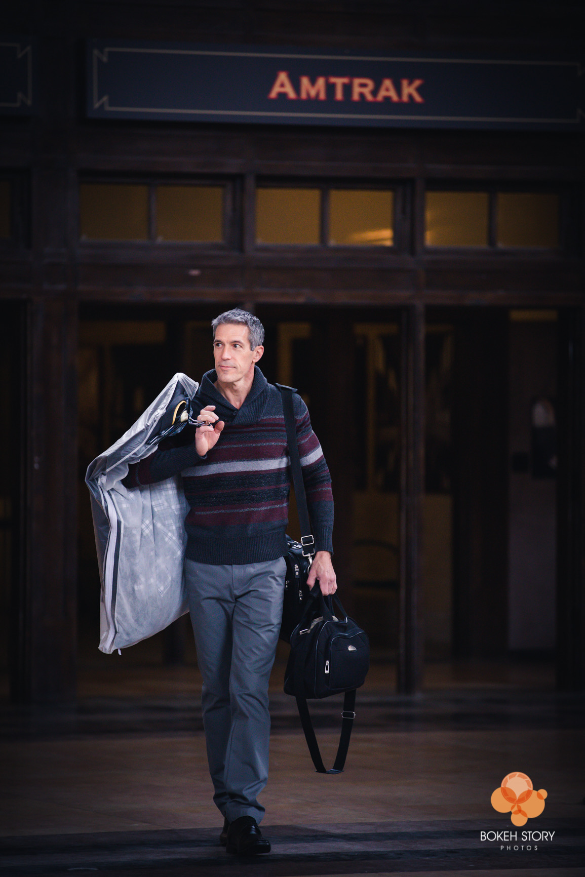 Portfolio portrait of a man walking through a historic train station carrying a garment bag and duffel bag, Amtrak signage visible behind him.