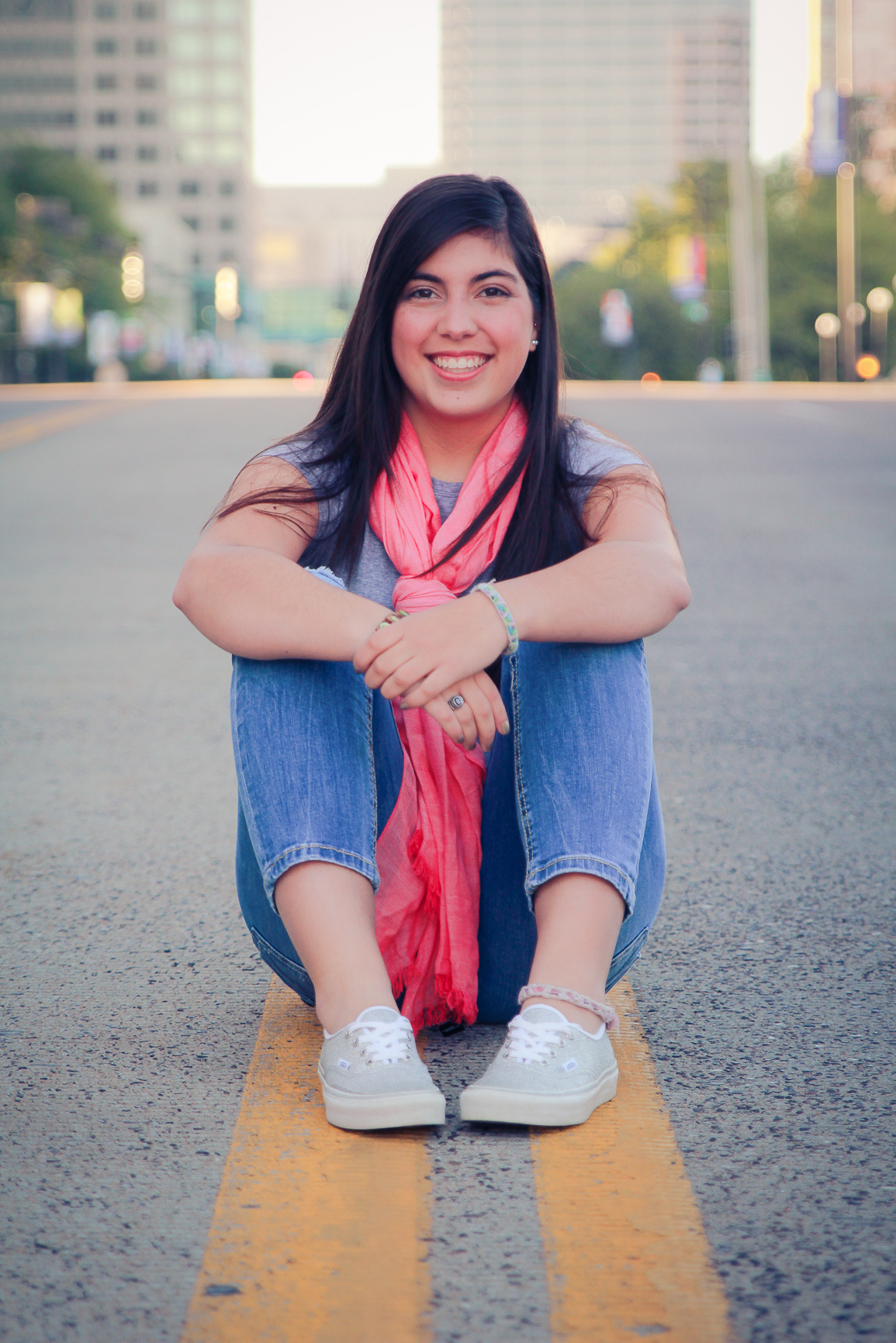 Senior portrait of a young woman sitting cross-legged on a city street, smiling at the camera.
