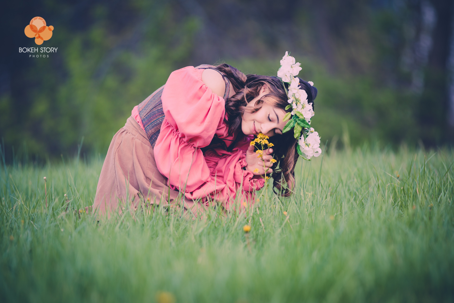 Senior portrait of a young woman in a flowing pink dress and flower crown, kneeling in tall grass to smell wildflowers.